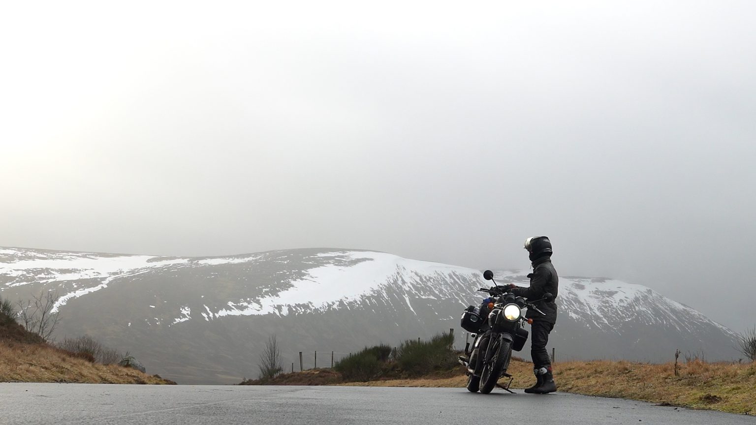 A rider and a Triumph Bonneville below a snowy Scottish hillside