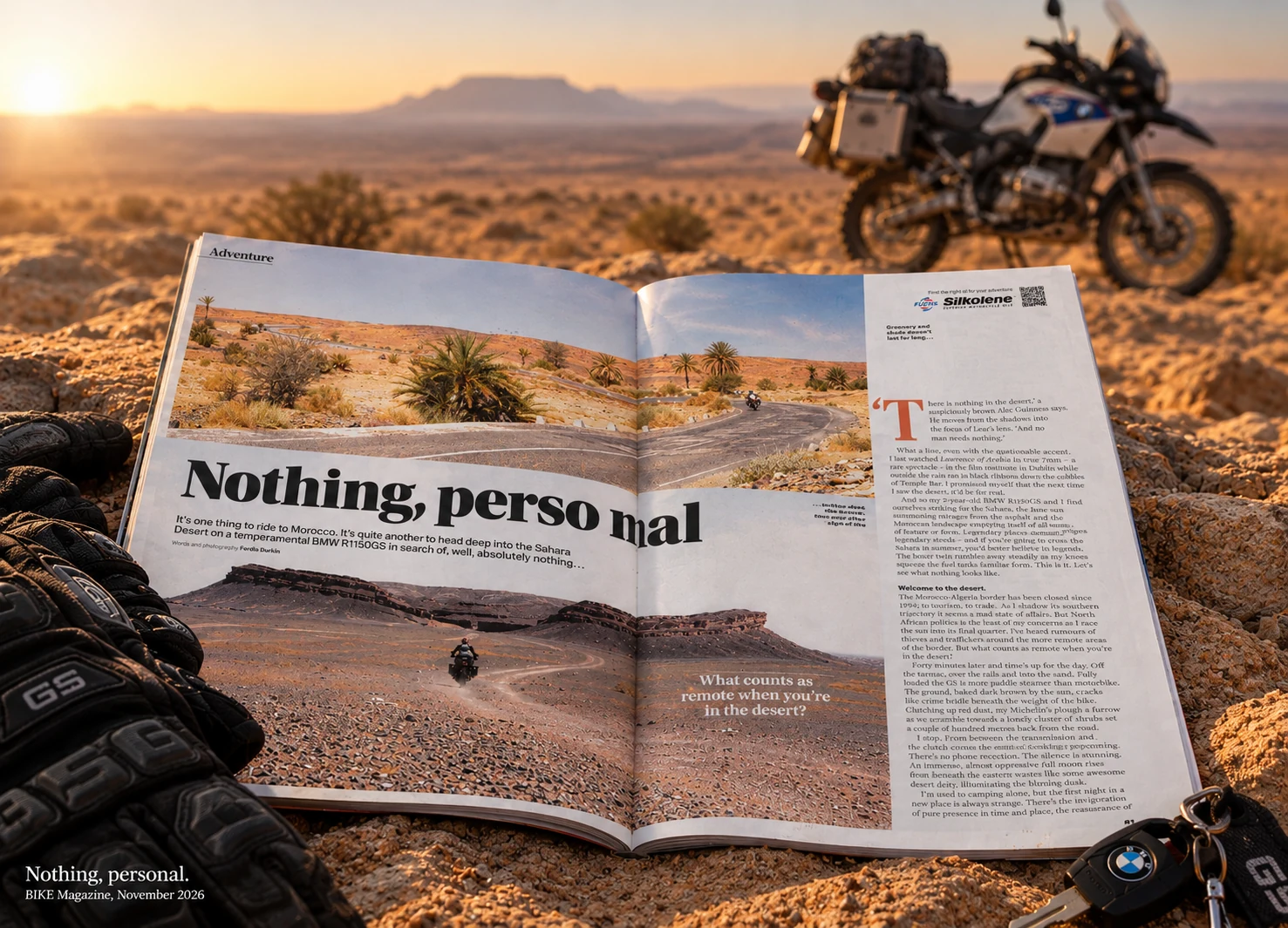 A copy of BIKE magazine open on desert sand at the Nothing, Personal spread, with riding gloves and a BMW key beside it and a loaded BMW GS out of focus in the sunset desert behind